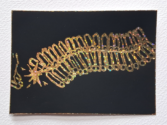 Une carte représentant une "mitraillette", un assemblage de 40 pétards reliés par une mèche centrale, évoquant un peu un mille-pattes, avec le bout allumé et une allumette en feu tenue par une main s'en éloignant ; gratté en holographique sur fond noir.