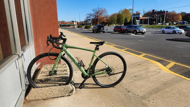 A green gravel bike with its front wheel parked in a poured concrete bike rack, set on a sidewalk next to a giant parking lot 