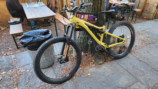 A yellow mountain bike with black fork. An assist motor is  attached to the crank. Bike is leaning against a tree, behind which is the outdoor dining setup of a cafe.