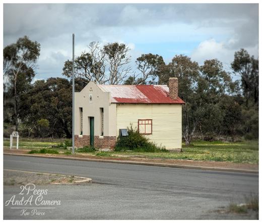 A small, historic looking building in Dumbleyung, Western Australia, with a distinctive design.
The front section is pale grey white with a small brick base and a peaked roof, while the main body is yellow cream corrugated metal with a rusty red corrugated iron roof and a brick chimney.
It sits near a paved road with a light post, surrounded by dry grass and scattered trees under a dramatic, cloudy sky.
