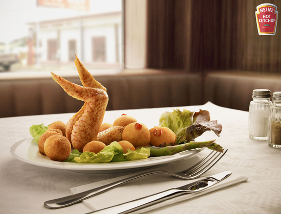 still life photograph of a plate of food set on a white table. The food consists of several fried potato balls arranged around a single, larger, fried potato shaped like a bird, with its wings outstretched. The plate also contains several small orange spheres, similar in size to cherry tomatoes, and a few pieces of green lettuce. To the right of the plate are two pieces of silverware – a fork and a knife – and two clear glass spice containers with white lids, one on either side. The background…