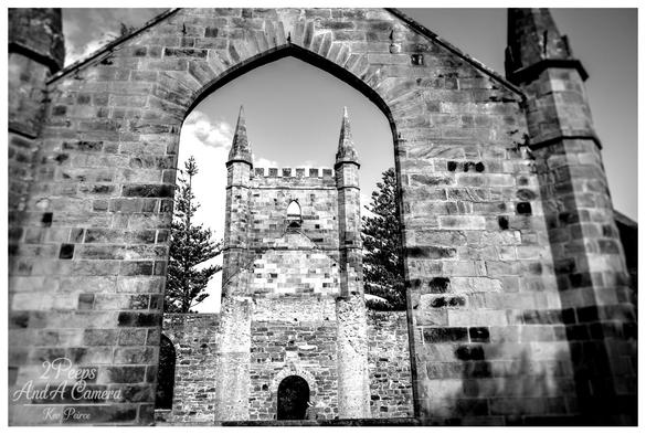 A high contrast, black and white photograph of the ruins of the Port Arthur Church in Tasmania. The shot is taken from inside the main structure, looking out through the large, arched main entrance.

This massive stone arch perfectly frames the central, tower like section of the church's ruins, which features pointed turrets and small arched openings.

Dark pine trees flank the central ruin, contrasting with the bright sky seen through the archway.