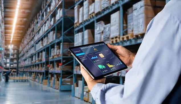 A warehouse manager using a digital tablet to monitor inventory and analytics in a large storage facility with tall racks filled with boxes, illustrating modern warehouse management technology.