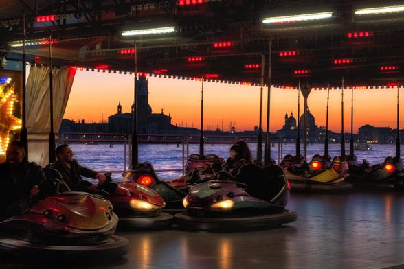 Farbfotografie, aufgenommen in der Dämmerung, die eine Autoscooter-Fahrbahn im Vordergrund mit einer berühmten Stadtkulisse am Wasser im Hintergrund kombiniert.

Im Vordergrund ist der innere Bereich einer Autoscooter-Anlage zu sehen. Die Decke ist mit Reihen von roten und weissen Neonröhren beleuchtet. Mehrere Personen sitzen in den glänzenden, dunklen Autoscootern, deren Scheinwerfer leuchten. Die Szene ist von dunklen, metallenen Haltestangen durchzogen.

Der Autoscooter-Bereich öffnet sich nach hinten zu einer Wasserfläche, über die die Stadtsilhouette von Venedig erstreckt.

Der Himmel bildet einen starken Kontrast: Er leuchtet in den kräftigen Tönen eines Sonnenuntergangs, von leuchtendem Orange und Gold nahe dem Horizont bis zu dunklerem Blau und Lila nach oben hin. Das tiefe Blau des Wassers trennt die beiden Ebenen.