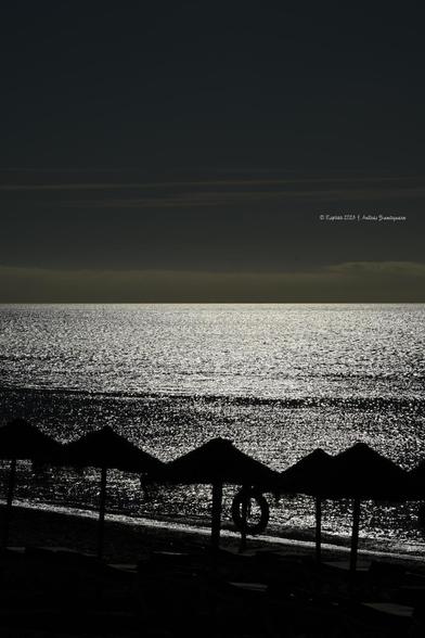 Imagen en formato vertical de unas sombrilla de playa junto a la orilla del mar fotografiadas en contraluz. El mar se ve lleno de destellos de luz solar y el cielo oscuro. Es una captura hecha por la mañana que el fuerte contraluz y algún que otro parámetro fotográfico hace que parezca que está oscuro. La imagen está dividida en dos partes casi simétricas: la mitad inferior son las sombrillas y mar con destellos y la superior es un cielo oscuro. En el límite se aprecian algunas nubes.
