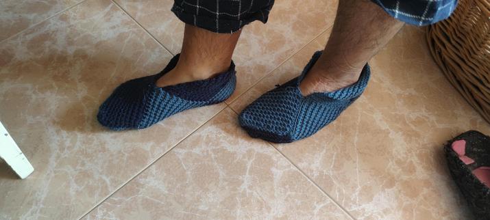 Persona usando pantuflas tejidas a mano en tonos azul oscuro y celeste, de pie sobre un suelo de baldosas color beige.