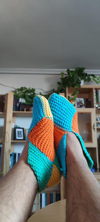 Un par de pies en pantuflas de ganchillo multicolores: naranja, amarillo, azul y turquesa.
El pie izquierdo se encuentra apoyado sobre el pie derecho, que está de frente.