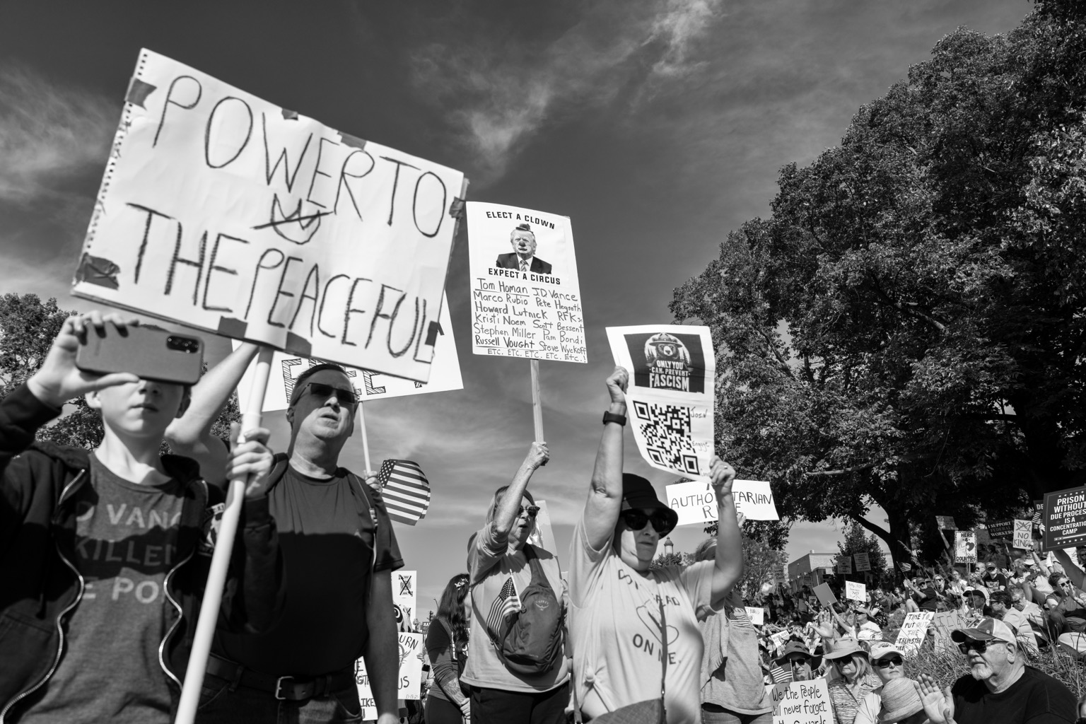 Black and white image of a No Kings Rally at the Iowa State Capitol in Des Moines, Iowa. A sign in the crowd of people can be seen reading, POWER TO THE PEACEFUL. 