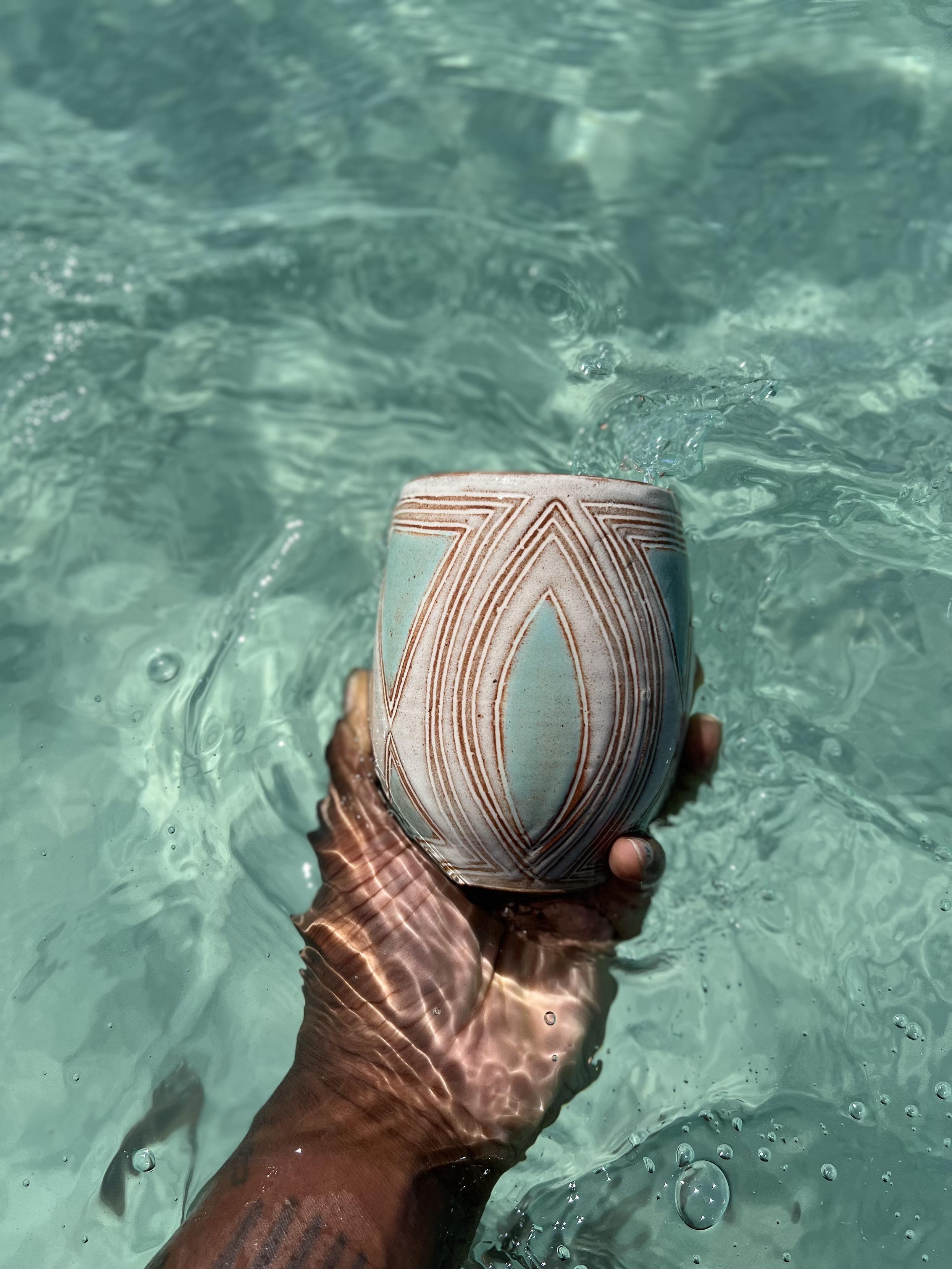 My hand holds a decorative ceramic cup above the surface of clear turquoise water, with light reflections and bubbles visible in the water. The cup is off white with carved geometric leaf-shaped patterns, with aqua color accents that match the water.