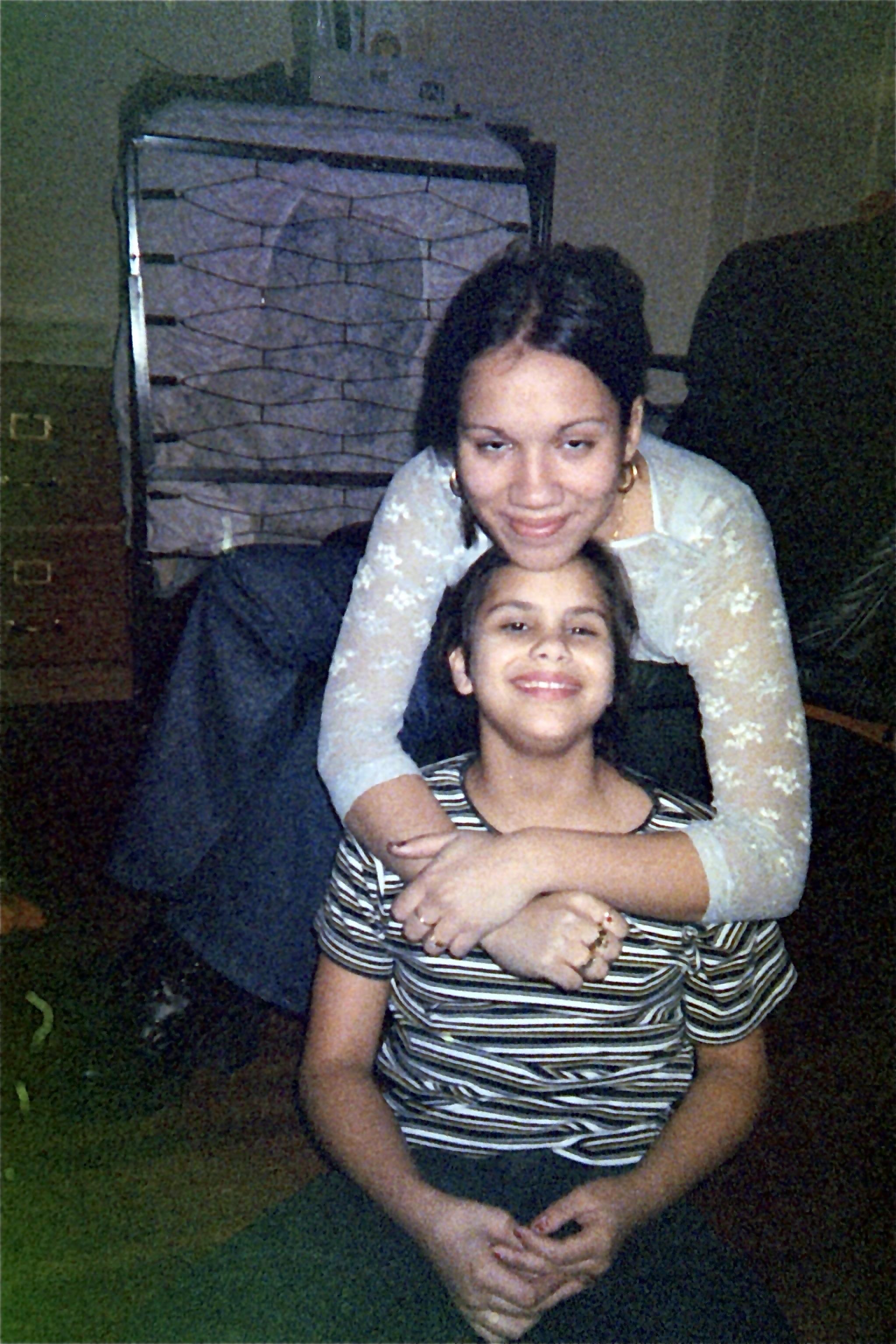 Two girls pose for a photo. One girl sits on the floor, smiling, while the other girl kneels behind her, embracing her from above. The background features a dresser and a bed. The lighting is warm and slightly soft.