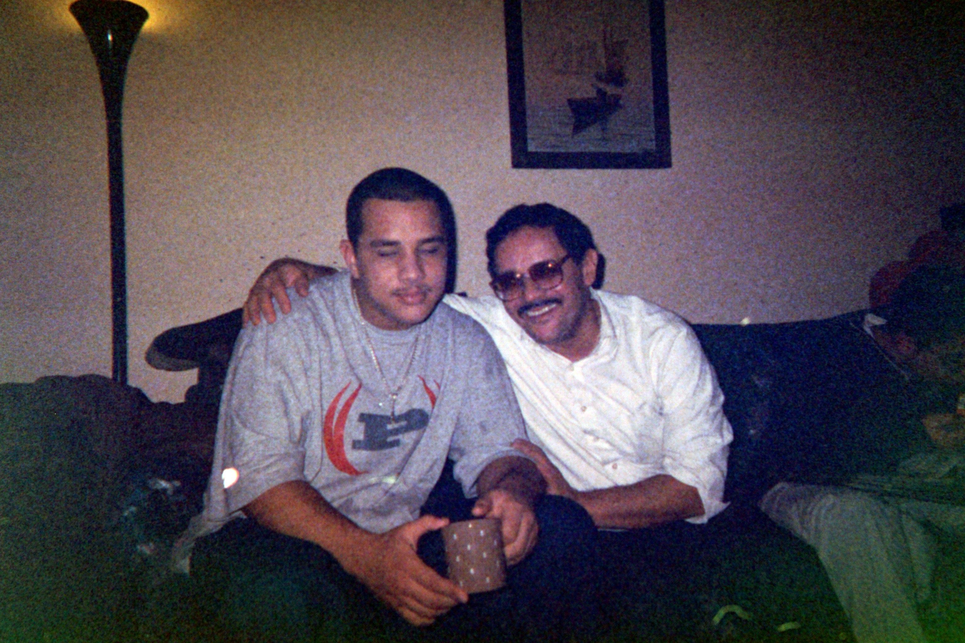 Two men smile while sitting together on a couch. One wears a gray t-shirt and holds a mug, while the other is in a white shirt and glasses. A dimly lit room with a lamp can be seen in the background.