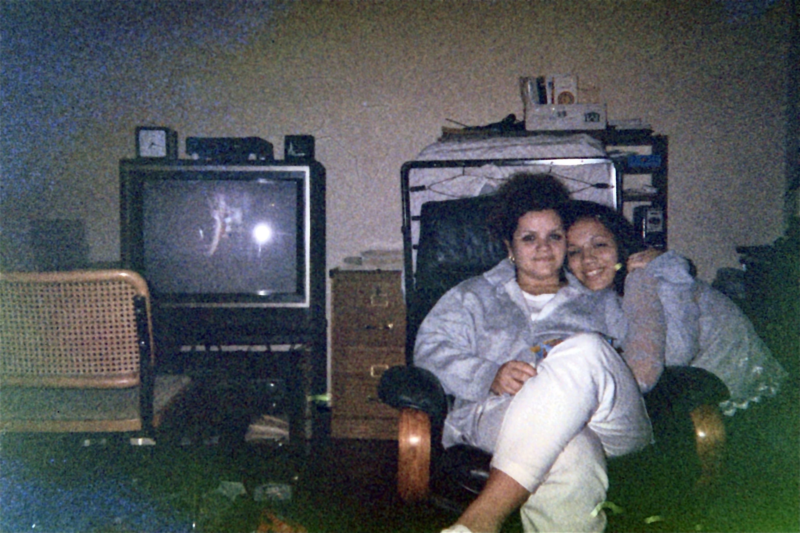 Two women sit closely together on a dark leather chair in a dimly lit room. One woman has curly hair and wears a gray jacket, while the other has straight hair and wears a blouse. A vintage television is visible in the background. 