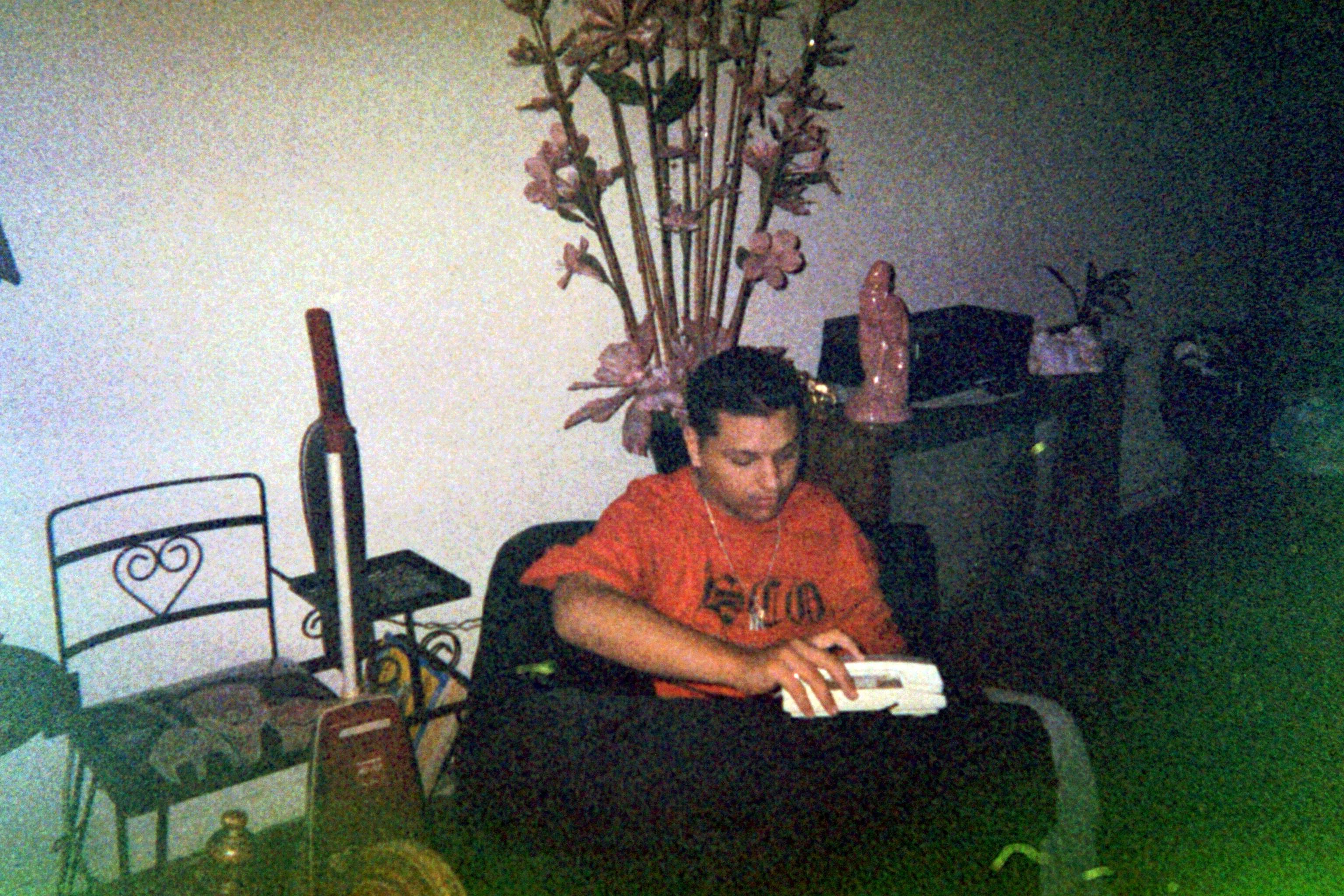 A young man in an orange shirt sits on a couch, focused on something in his hand. There is a decorative plant in the background, alongside various household items and a statue. The lighting is dim, giving the image a retro feel.