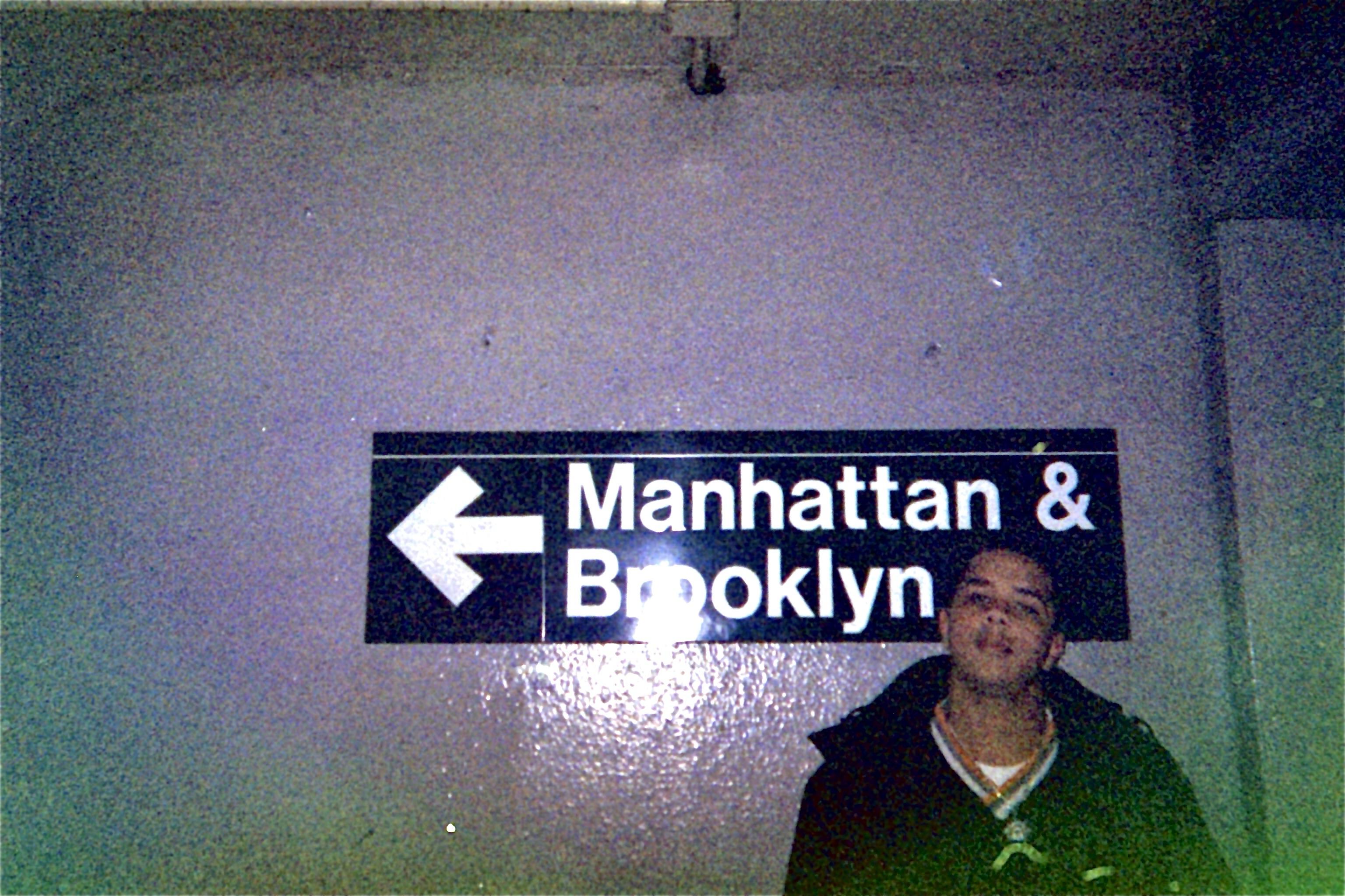 An indoor wall sign indicating directions to Manhattan and Brooklyn, with a person standing beside it. The sign features an arrow pointing left.