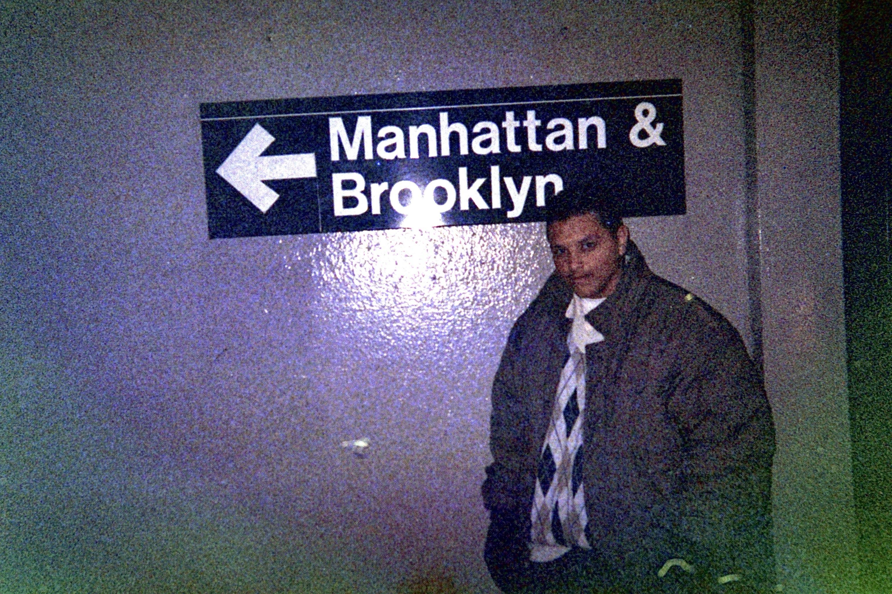 A person stands next to a directional sign indicating "Manhattan & Brooklyn." The setting appears to be an underground or urban area, with dim lighting.