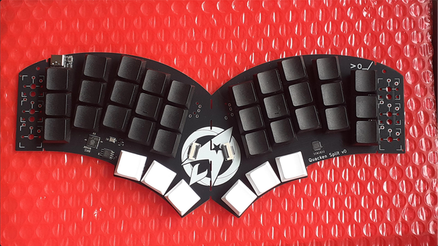 Photo d’un clavier ergonomique noir, posé sur un bullback rouge.
Le clavier est un monobloc splittable,  monté en 3×5 touches par main (noires) + 3 touches par pouce (blanche). On voit que les 6e colonnes, non équippées, sont sécables. Au centre, un logo Ergo‑L occupe la place du touchpad circulaire qui équipera la version suivante.