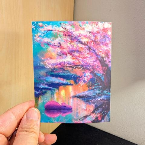 Photo of a left hand holding a rectangle sticker. The sticker is a matte painting of a capybara resting under a cherry blossom tree.