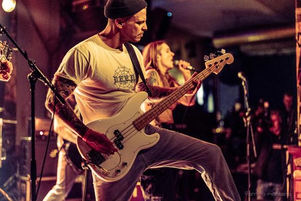 A bassist performs on stage, wearing a white t-shirt and a beanie, with a focused expression. A female vocalist is singing in the background. The setting is a live music venue, illuminated with warm lighting, capturing the energy of the performance.