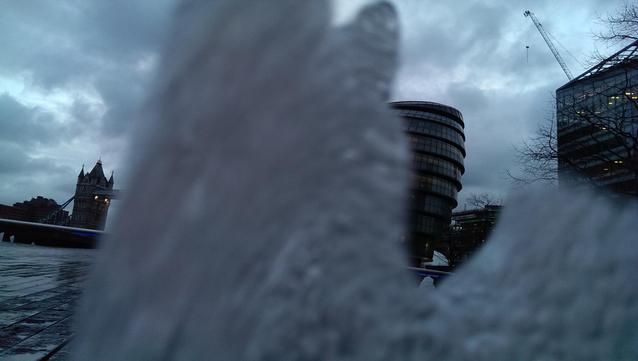 A close up of a foam of water from a soaked floor fountain almost obstructs the whole frame of this early morning photo of a gherkin shaped building on the right and a part of a tower bridge on the left against a dull cloudy sky just before sunset. 
The other three photos are a variation of the first but the buildings and other  surroundings are more visible.