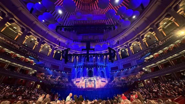 Wide angle view of the Royal Albert Hall stage and ceiling, lit up in purple and red. The audience are getting ready for a concert