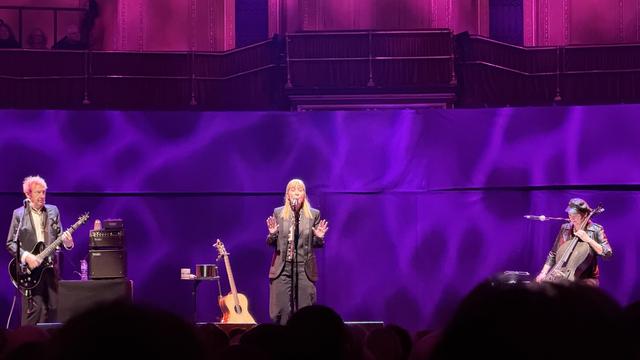 Suzanne Vega performing at the Royal Albert Hall. She’s singing into the mic, palms forward, with a purple water reflection pattern in the background. 