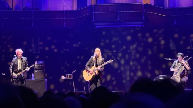 Suzanne Vega’s band. She’s singing and playing acoustic guitar. To her left is Gerry Leonard on electric guitar, with Stephanie Winters on cello to her right. 
