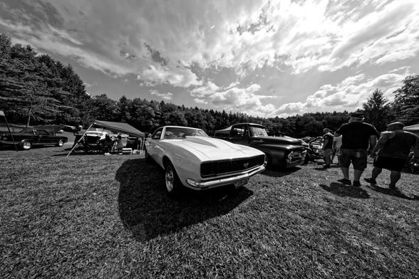 White first-generation Chevy Camaro SS is in the center and kinda small compared to an amazing wide angle sky and other cars, spectators and grass.