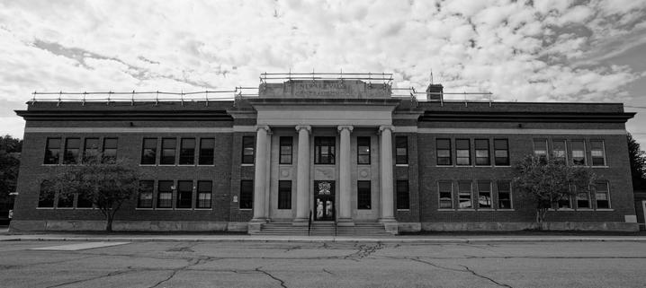Distinguished old school building with classical columns in the center and rows of windows in brick to the sides with a cracked parking lot in front and mackerel sky above 