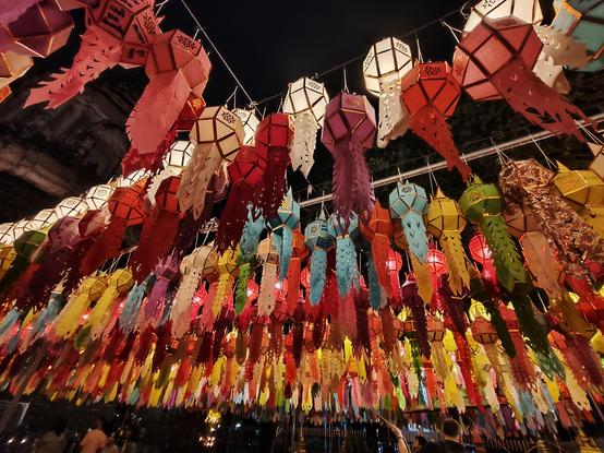 This close-up photo shows hundreds of colorful Lanna-style lanterns strung tightly overhead, creating a vibrant, glowing canopy. The lanterns are white, pink, red, blue, green, and yellow, with long, tasseled tails dangling down toward the crowd below.

