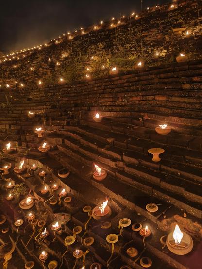 This dramatic photo features a large, sloping brick wall, likely a temple structure, densely decorated with small, flickering oil lamps. The numerous terracotta lamps are placed on the stepped ledges of the dark brickwork, creating a warm, glowing pattern of firelight that ascends into the darkness.