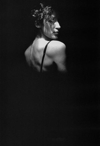 Black and white photograph of a person’s upper body and side profile, dramatically lit against a very dark background. The person is facing to the right, with a visible shoulder, arm, and neck. A strap crosses the back and extends over the shoulder, suggesting some sort of garment. The person has short, curly hair and a defined jawline and nose. The majority of the image is obscured by shadow, creating a high-contrast, moody effect.

Schwarz-Weiß-Foto des Oberkörpers und des Seitenprofils einer Person, dramatisch beleuchtet vor einem sehr dunklen Hintergrund. Die Person ist nach rechts gerichtet, mit sichtbarer Schulter, Arm und Nacken. Ein Riemen kreuzt den Rücken und erstreckt sich über die Schulter, was auf eine Art Kleidungsstück hindeutet. Die Person hat kurzes, lockiges Haar und eine definierte Kieferlinie und Nase. Der Großteil des Bildes wird durch Schatten verdeckt, wodurch ein kontrastreicher, launischer Effekt entsteht.