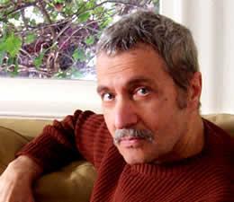 Profile image of author Michael Parenti sitting on a chair in front of an abstract painting. 