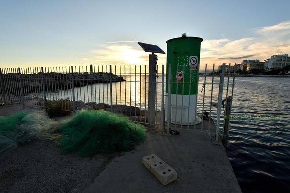 Imagen donde se ve la marca lateral de estribor de entrada al canal del puerto tras una valla. En primer término se ve una malla verde, una red, y un bloque de cemento con seis orificios, posiblemente para fijar postes y vallas. La tarde cae y el cielo tiene alguna nubosidad.