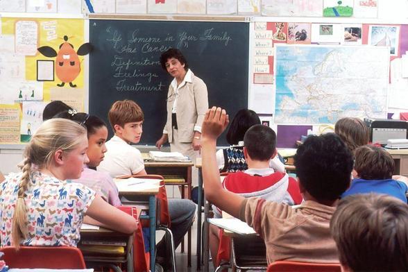 A teacher interacts with students in a classroom, where one student raises a hand to participate. The image represents learning and dialogue in education.