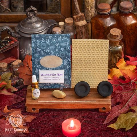 The contents of our exclusive "Gemini Full Moon Ritual Kit for December 4th" product are arranged on a small wood platform and surrounded by faux fall foliage. They include a sheet of yellow beeswax and a wick; a pouch of December Full Moon herbs, including chamomile, peppermint, lemon balm, and lemon peel; a dropper vial of our exclusive Full Moon Spell Oil; a tumbled serpentine gemstone; and two charcoal discs. Presented by Inked Goddess Creations.
