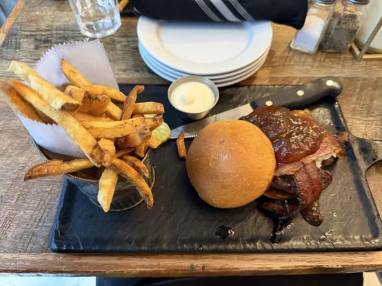 A plate featuring a burger topped with bacon and fig sauce, alongside a bun. A small container of dipping sauce and a serving of golden, crispy fries in a cup are also included. The meal is presented on a black slate surface.