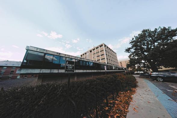 A roughly cubical building rises above a wide second story clad in glass on the left side of a sidewalk that runs up and down near the right of the frame with a big tree to the right