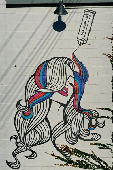 A mural of a woman is painted on a concrete block wall  with bands of color coming out of a tube that say HAIR * COLOR * ART