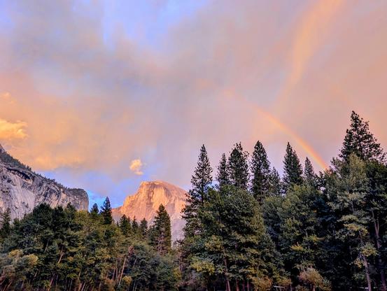 Rainbow over Half Dome at sunset. The clouds are tinted pink and salmon, with glimpses of brilliant blue sky behind them. Photograph taken by me during a recent camping trip in September. CC BY-SA Kate Zimmerman.