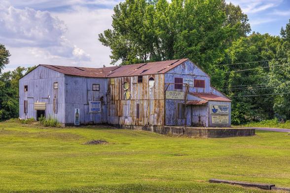 An old, weathered building with rusted metal siding sits on a grassy field, surrounded by lush green trees.