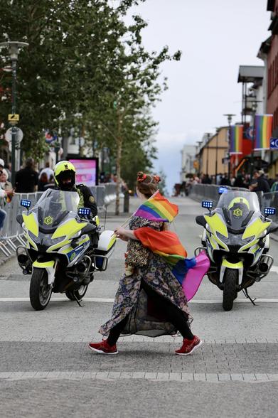 A woman wrapped in pride flags walks past two police motorcycles as one officer looks on. Reykjavík Pride 2024