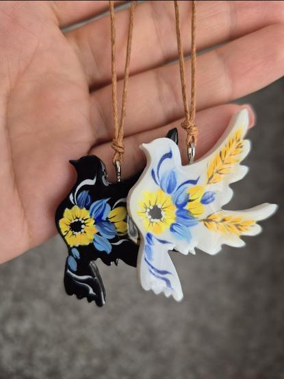 A hand holds two dove-shaped pendants, painted with Ukrainian blue-and-yellow flowers and a sunflower. One dove is black, the other is white.