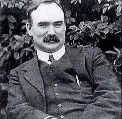 Old black and white profile photograph of James Connolly sitting casually, slightly reclined and smiling, in front of hedging. He is slightly smiling. 