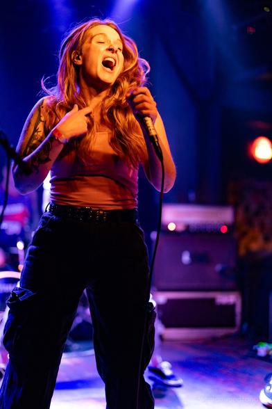 A female singer performs on stage, passionately engaging with the audience. She has long hair and a nose ring, and wears a tank top paired with cargo pants. The background features soft stage lighting, creating a vibrant atmosphere.