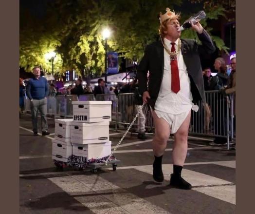 Image of a man dressed like Trump walking down the street pulling a wagon with 5 while file boxes marked, “Epstein”.  The man is wearing a black blazer, white dress shirt, red tie, diaper, black socks and shoes, as well as a crown and gold necklaces.  He is also drinking from a bottle.