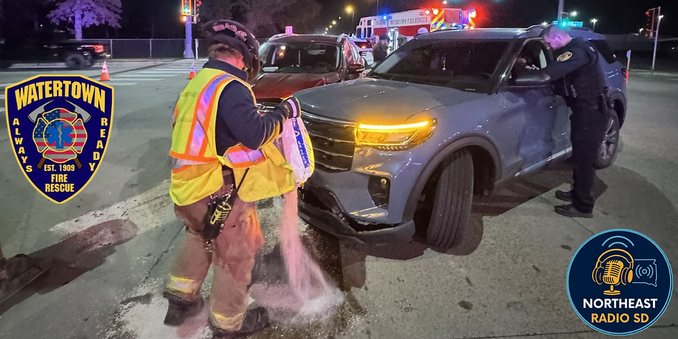 Firefighter pours absorbent near a car at night, with police officer talking to driver. Emergency vehicles and cones in background. Watertown Fire Rescue logo visible.