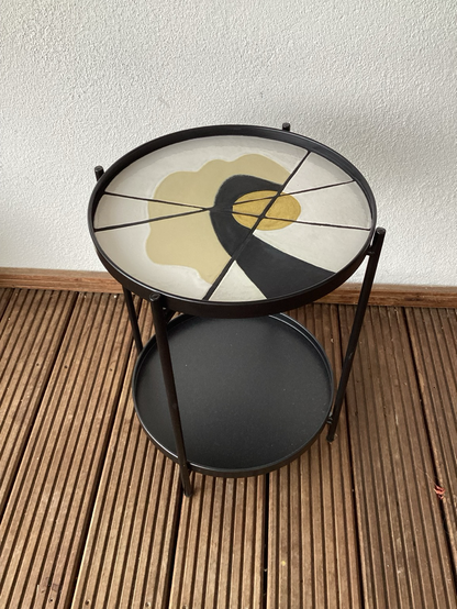 Schwarzer, runder Beistelltisch mit Keramikfliesen  (gelber Kreis, teilweise umgeben von einem schwarzen Bogen, einer beigen Wellenlinie und einer weißen Restfläche. Die unterschiedlich geformten Teilsegmente sind mit schwarzen Fugen verbunden.