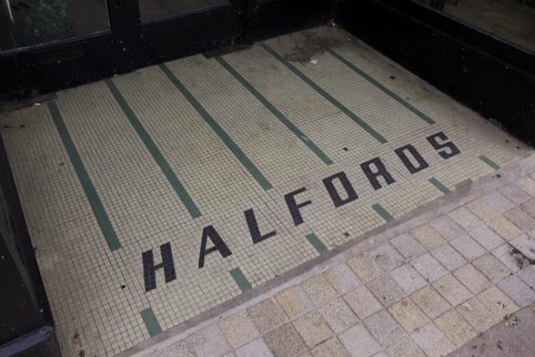 Mosaic tile entrance with the word “HALFORDS” spelt in black tiles. The background is light-coloured with green vertical lines, leading to black-framed glass doors. Adjacent pavement consists of square beige and tan tiles.