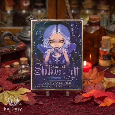 A boxed deck of oracle cards stands on a red lace-covered surface. Its front features a beautiful stylized fairy girl with purple hair curling around her face and purple butterfly wings. She has doll-like proportions with a large head and eyes, which look tear-filled. The deck's box is labeled "Sun-Steeped / a golden hour-infused affirmation deck / Shannon Christie". Presented by Inked Goddess Creations.