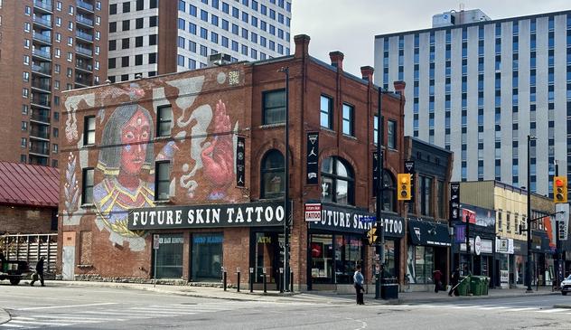 Future Skin Tattoo Laden.
Roter dreigeschossiger Backsteinbau zwischen Hochhäusern.
Ein ganzflächiges Mural bedeckt eine Seitenwand.
